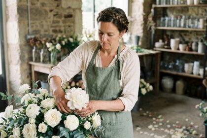 Fleuriste événementielle parisienne arrangeant un centre de table nuptial avec pivoines blanches et roses dans son atelier artisanal